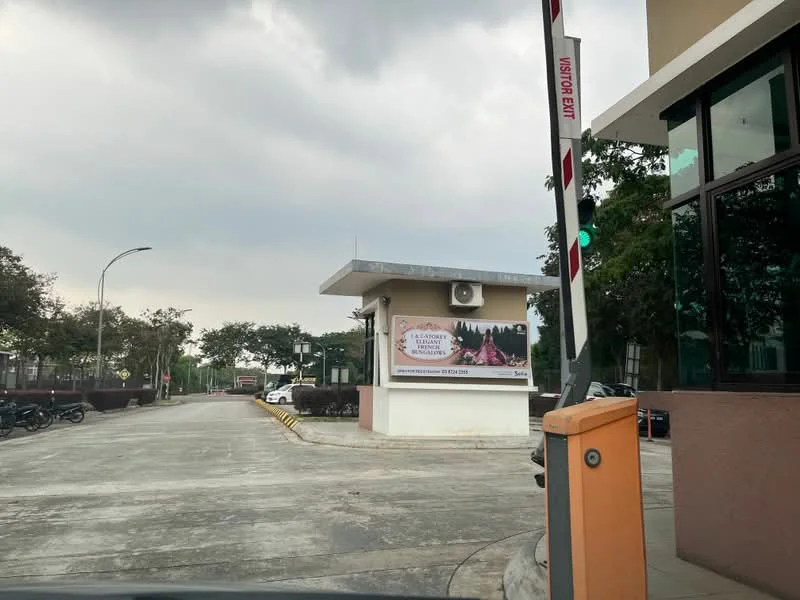 Exterior type: main entrance gate; features: guard house at entrance, boom gate, CCTV cameras visible, perimeter fence, landscaping with trees, paved driveway, advertisement banner, parking area for vehicles; style: modern.