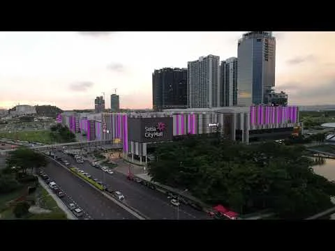 Exterior type: shopping mall; features: modern facade with colorful lighting, multiple storeys, large glass windows, surrounded by greenery, adjacent to a main road, parking area visible, urban context with high-rise buildings in background; style: contemporary.