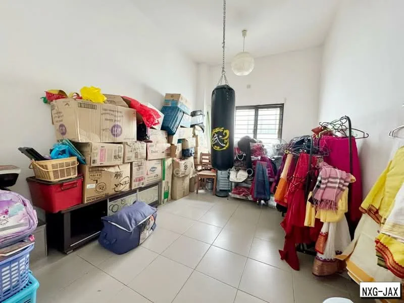 Room features: white painted walls, tile flooring, large window, boxing bag, stacked cardboard boxes, clothing rack with garments, various bags and items on the floor, suspended light fixture.