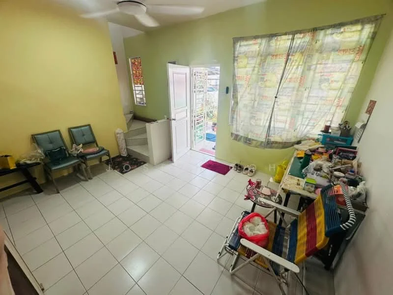 Room features: light green painted walls, white tiled flooring, ceiling fan, large window with sheer curtains, staircase visible, two green chairs, folding chair, small table, various items on the floor, door leading outside.