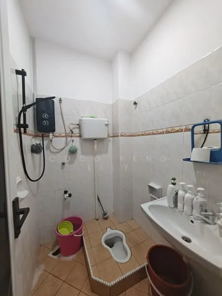 Bathroom features: squat toilet, wall-mounted sink, chrome faucet, white tiled walls, beige tiled flooring, toilet paper holder, soap dispenser, cleaning supplies, bucket, shower hose, wall-mounted mirror, towel rack.