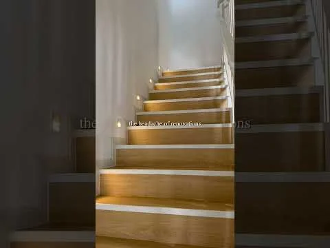Room features: wooden staircase, light-colored wood steps, white painted handrail, recessed lighting along the staircase, neutral wall color, minimalist design; style: modern.
