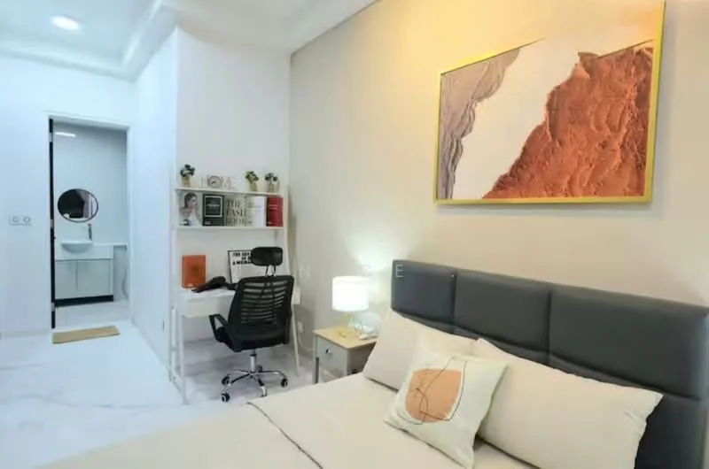 Bedroom features: king-size bed with grey upholstered headboard, white bedding with decorative pillows, nightstand with lamp, desk with chair, wall-mounted artwork, marble flooring, built-in wardrobe, recessed ceiling lights, floor-to-ceiling mirror, white painted walls; style: modern. Bathroom features: vanity with sink, mirror above vanity, white tiled walls, toilet, shower area, light fixture, towel rack.