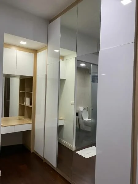 Room features: built-in wardrobe with mirrored doors, white glossy cabinetry, wooden countertop, vanity area with mirror, recessed lighting, dark wood flooring, neutral wall colour, toilet visible in reflection; style: modern.