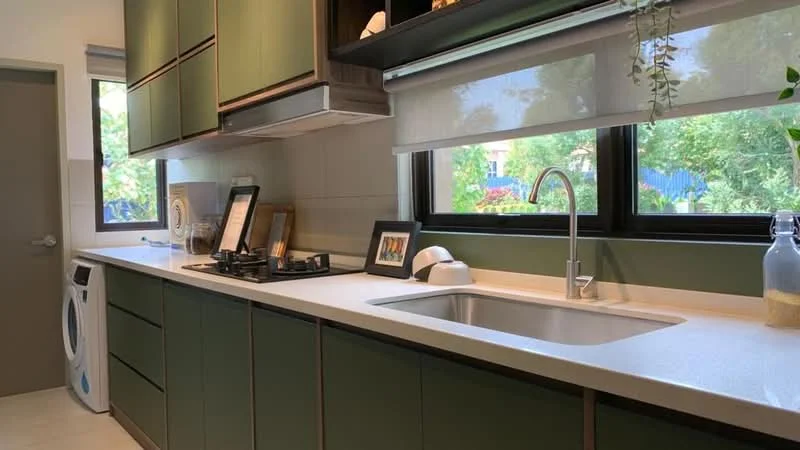 Kitchen features: green cabinetry with flat-panel design, white quartz countertop, single-bowl stainless-steel sink, chrome pull-down faucet, built-in gas stove with two burners, white washing machine, large windows with garden view, roller blinds, decorative plants on windowsill, framed artwork on countertop; style: modern; view: garden.