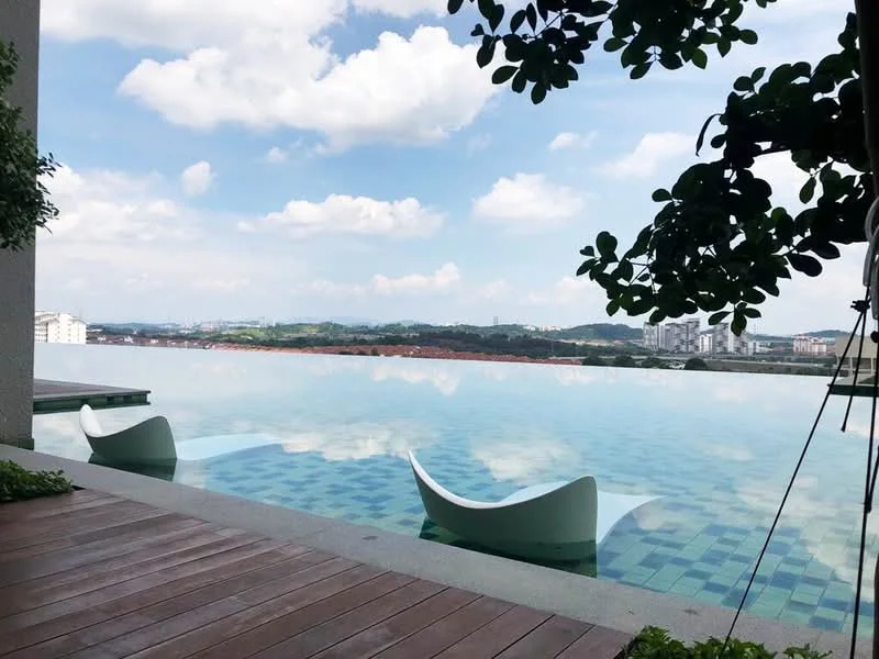 Exterior type: swimming pool deck; features: infinity pool, pool deck with loungers, landscaped greenery, city skyline in background, blue mosaic tile flooring, surrounded by trees, clear blue sky, clouds reflecting on water; style: modern.