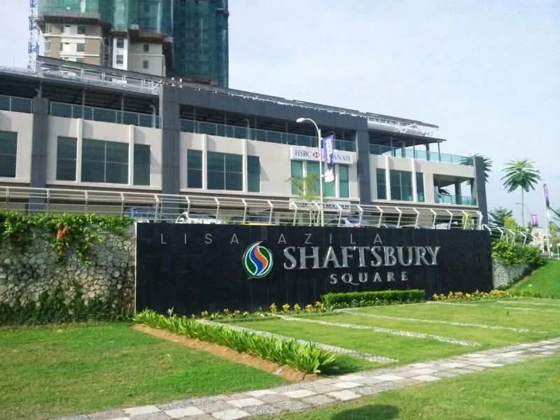 Exterior type: commercial building; features: glass facade, signage with 'SHAFTSBURY SQUARE', manicured lawn, stone-paved walkway, landscaped garden with plants, multi-storey structure, visible construction scaffolding, parking area, street lighting; style: modern.