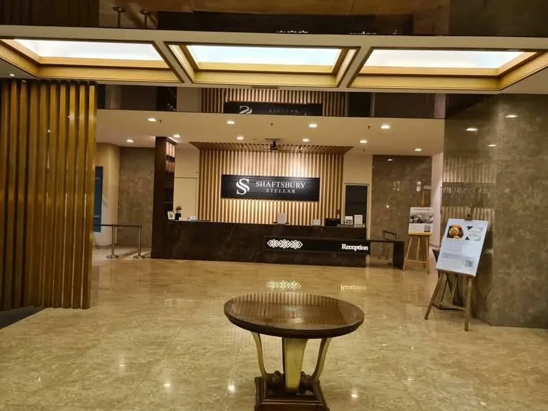 Lobby features: marble flooring, wood paneling on walls, reception desk with logo, backlit feature wall, ceiling with recessed lighting, hanging light fixtures, decorative wall art, seating area with chairs, information stand, large windows, open layout; style: modern.