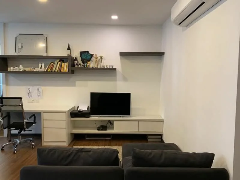 Living features: grey fabric sofa, TV console, wall-mounted TV, floating shelf, decorative items on shelf, white painted walls, wooden flooring, 1 wall-mounted split AC, glass office chair, desk with drawers; style: modern.