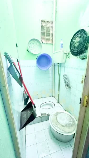Bathroom features: white tiled walls, light green painted walls, white ceramic toilet, floor-mounted toilet, white tiled flooring, sink with faucet, cleaning supplies on wall, hanging bowls, window for natural light, broom and mop, shower area with no enclosure.
