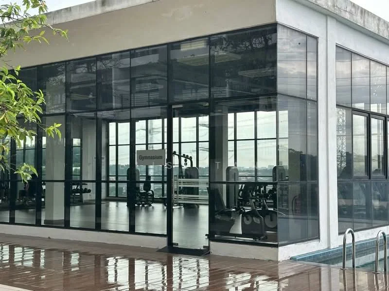 Exterior type: gymnasium; features: glass facade, metal framing, large windows, outdoor swimming pool, reflected surface on the ground, surrounding greenery, modern design, open layout, visible gym equipment inside; style: modern.
