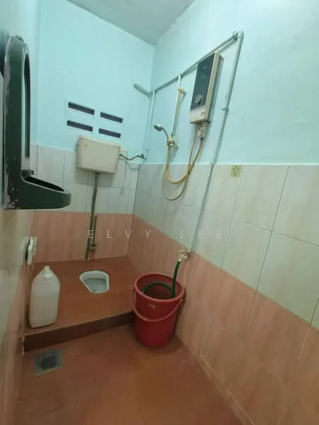 Bathroom features: squat toilet, wall-mounted shower head, water heater, sink with tap, ceramic tile flooring, pink tiled walls, drainage grate, plastic bucket, cleaning liquid container.