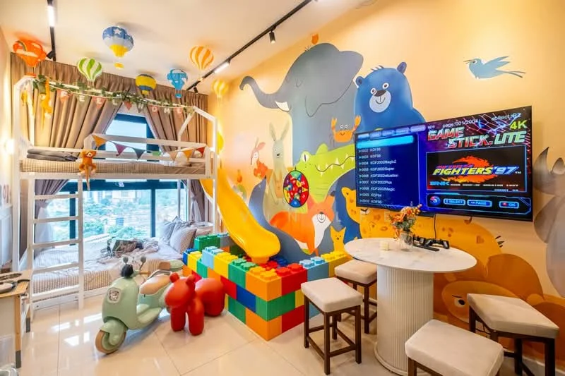 Room features: bunk bed with slide, colorful animal-themed wall mural, large flat-screen TV, gaming console, round dining table, stools, colorful building blocks, decorative balloons, large windows with city view, light-colored tiled flooring, curtains, playful decor elements; style: modern; view: city.