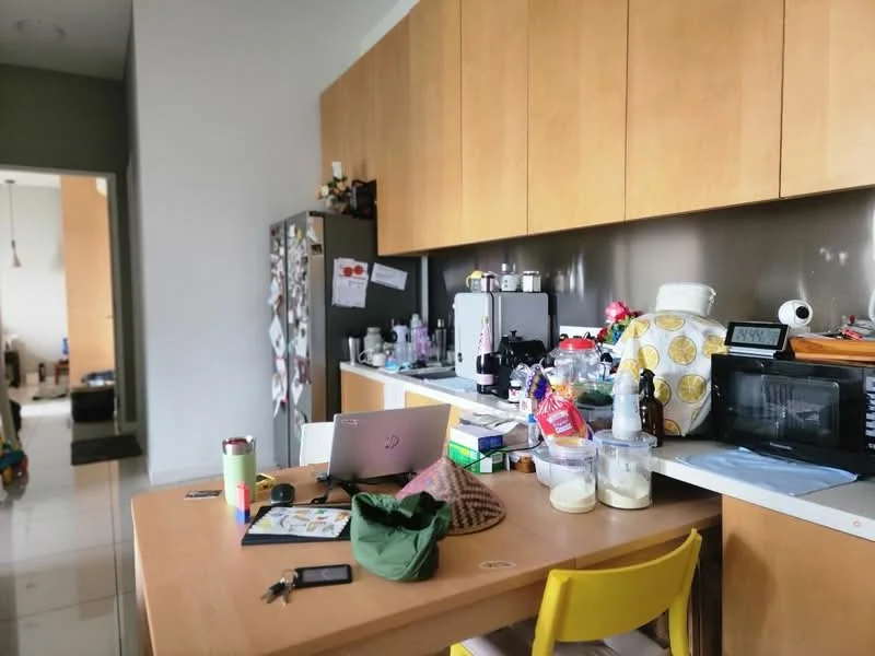 Kitchen features: wood cabinetry in light brown, white countertop, stainless-steel fridge, microwave, black countertop oven, kettle, blender, various kitchen utensils, table with yellow chairs, laptop on table, decorative items on countertop, tile flooring, grey accent wall; style: modern.