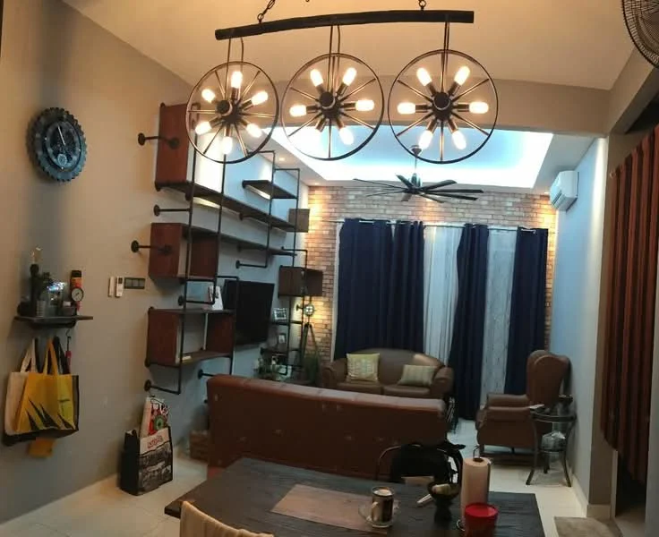 Living features: 3-blade ceiling fan, chandelier with exposed bulbs, grey painted walls, exposed brick accent wall, dark wood shelving units, brown leather sofa, armchair in brown leather, dark curtains, coffee table, decorative wall clock, area rug, wall-mounted TV, decorative plants, open shelving with decorative items; style: industrial.
