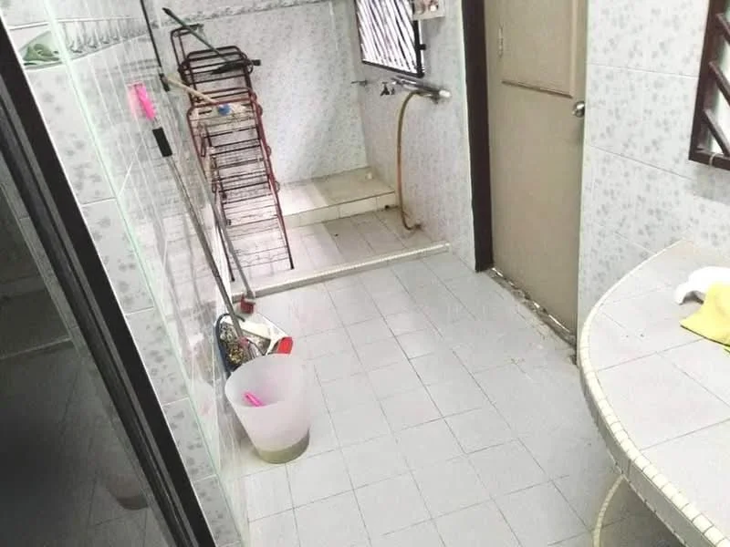 Bathroom features: white tiled walls, light grey tiled flooring, single-sink vanity with circular basin, mirror above vanity, shower area with no enclosure, clothes drying rack, toilet, plastic bucket, drainage pipe visible, door leading to another room.