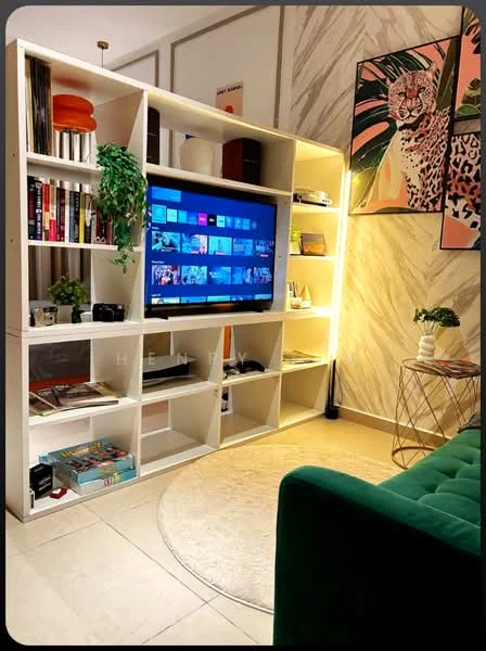 Living features: green velvet sofa, round area rug, white open shelving unit, wall-mounted TV, decorative plants, framed artwork, books on shelves, LED strip lighting, coffee table, decorative items on shelves; style: modern.