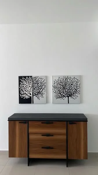 Room features: wooden sideboard with three drawers, black countertop, two framed artworks with tree designs in black and white, white painted walls, light-colored tile flooring; style: modern.