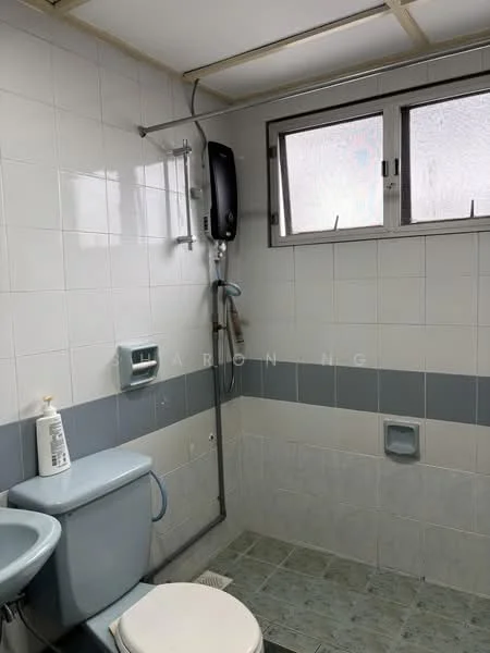 Bathroom features: white tiled walls, grey tiled accent strip, light green ceramic tile flooring, toilet, single-sink vanity, shower area with glass partition, handheld shower, water heater, window for natural light, wall-mounted soap dispenser.