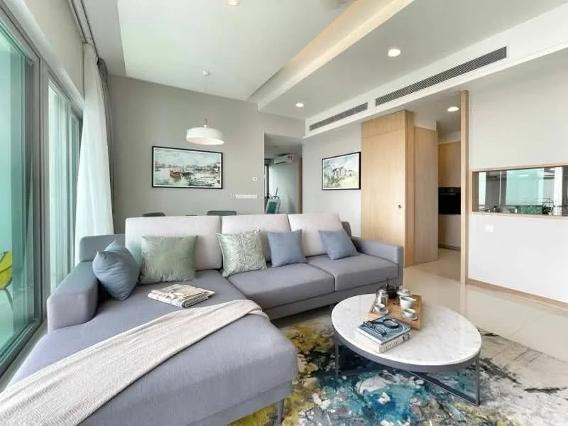 Living features: L-shape sectional sofa in grey, decorative cushions in various colors, round coffee table with marble top, area rug with abstract pattern, pendant light fixture, floor-to-ceiling windows, curtains, wall-mounted artwork, light-colored walls, recessed ceiling lights, open layout to kitchen, wooden cabinetry, neutral-colored flooring; style: modern; view: city.