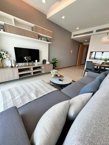 Living features: grey fabric sectional sofa, decorative throw pillows, round coffee table, wall-mounted TV, light wood TV console, white shelving unit, grey accent wall, recessed ceiling lights, dining area visible in background, large windows with natural light; style: modern.