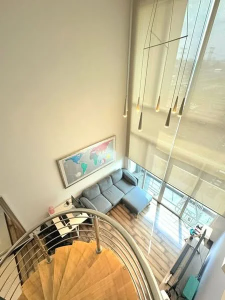 Living features: grey fabric sectional sofa, wooden flooring, large windows with sheer blinds, pendant lighting fixtures, framed world map artwork, open layout, spiral staircase, high ceiling; style: modern; view: city.