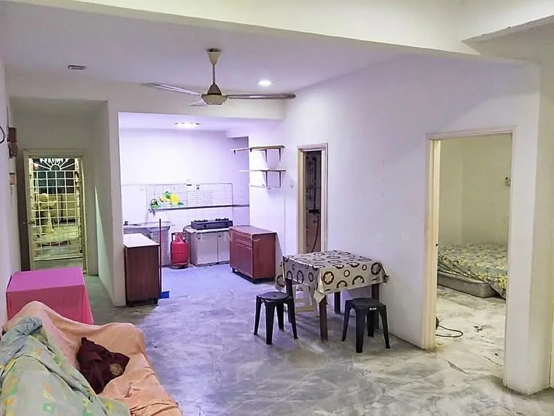 Living features: sofa in light fabric, ceiling fan, dining table with patterned tablecloth, 4 black plastic chairs, grey tiled flooring, white painted walls, open layout to kitchen, natural light from doorway. Kitchen features: kitchen counter with sink, gas stove, wooden cabinetry, red gas cylinder, open shelving, grey tiled flooring. Bedroom features: bed with mattress, white painted walls, grey tiled flooring, doorway to hallway.