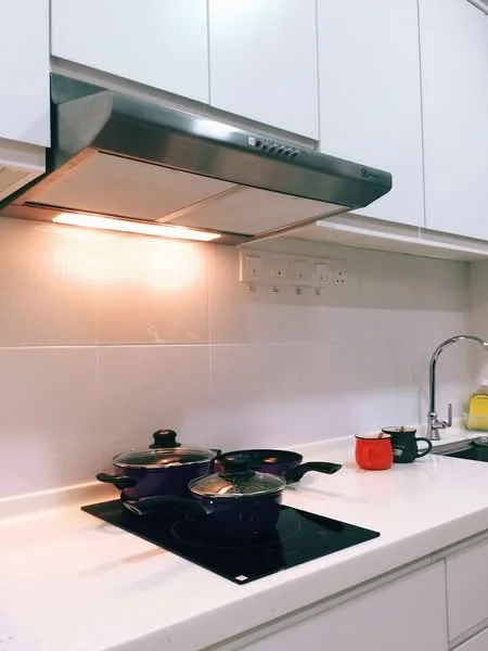 Kitchen features: white cabinetry, stainless-steel range hood, ceramic tile backsplash, white countertop, black induction cooktop, purple cooking pots, red kettle, yellow dish soap, under-cabinet lighting; style: modern.