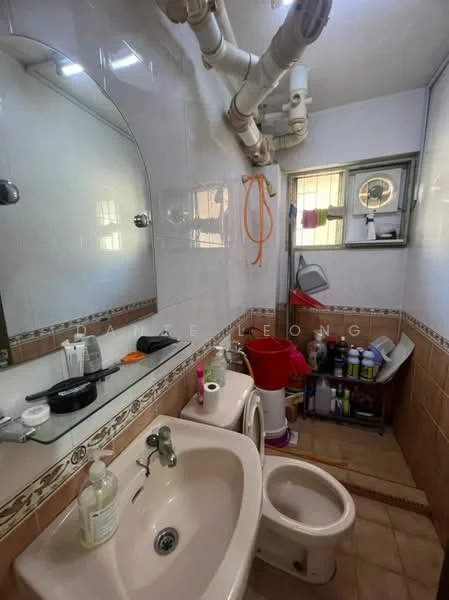 Bathroom features: white ceramic sink, chrome faucet, mirror above sink, white toilet, tile flooring, white tiled walls, brown tiled accents, window with security bars, visible plumbing pipes, shelf with toiletries, cleaning supplies in storage, hanging towel rack.