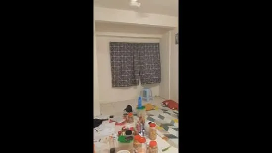 Room features: white painted walls, ceiling fan, grey patterned curtains, various containers and items on the floor, multicolored patterned flooring, scattered papers and objects.