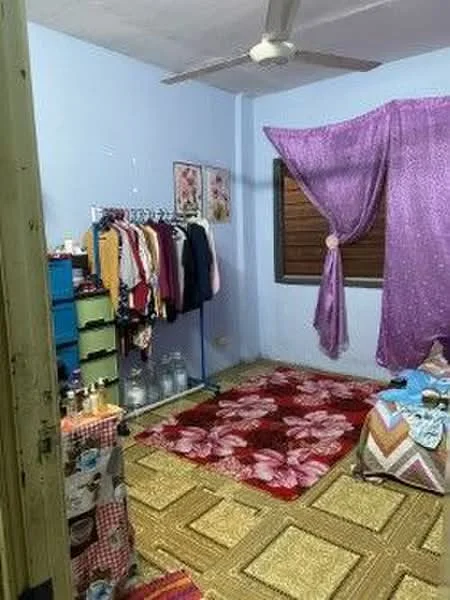 Bedroom features: light blue painted walls, ceiling fan, window with wooden blinds, purple curtain, red floral area rug, beige patterned flooring, clothing rack with assorted clothes, blue storage drawers, bed with colorful bedding, decorative wall art.