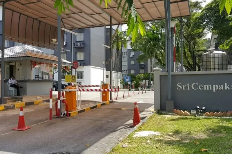 Exterior type: gated community; features: guard house at entrance, boom gate, CCTV cameras visible, perimeter fence, manicured lawn, paved driveway, signage with name 'Sri Cempaka', surrounded by greenery; style: modern.