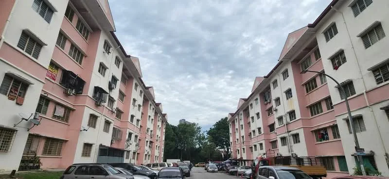 Exterior type: low-rise apartment block; features: pink and white painted facade, 3-storey low-rise, multiple windows with grilles, balconies with laundry hanging, visible air conditioning units, surrounded by greenery, open-air parking lot, street lighting, asphalt road, cloudy sky; style: modern.