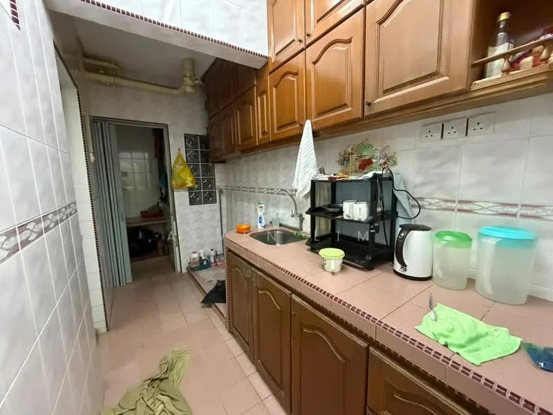 Kitchen features: brown wooden cabinetry, white tiled walls, pink tiled flooring, single-bowl stainless-steel sink, chrome pull-down faucet, black coffee machine, white kettle, clear plastic storage containers, green cloth on countertop, yellow plastic bag hanging, wall-mounted shelves, exposed pipes, doorway leading to another room.