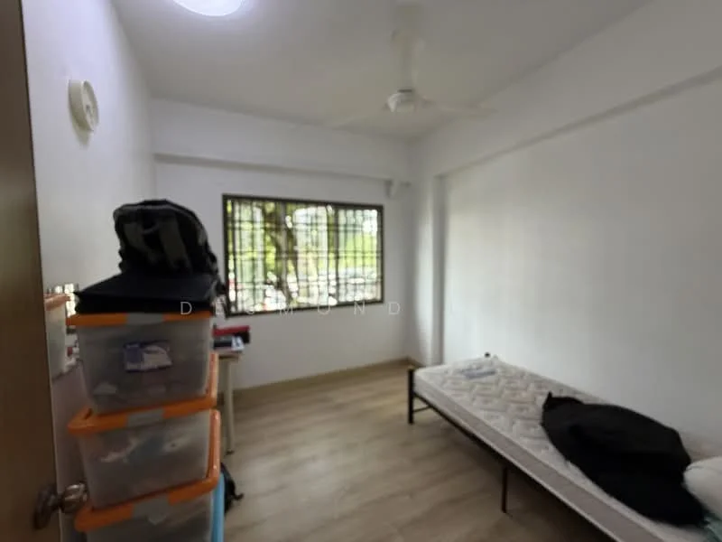 Room features: wooden flooring, white painted walls, ceiling fan, window with metal grille, single bed frame without mattress, plastic storage boxes, small table, black bag on the floor; view: garden.