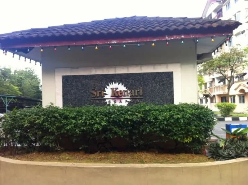 Exterior features: signage with the name 'Sri Kenari', stone cladding on the sign, decorative lighting, landscaped greenery, manicured bushes, paved walkway, surrounding residential buildings; style: modern.