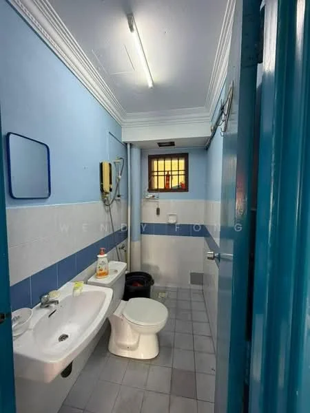 Bathroom features: white ceramic toilet, single-sink vanity with white cabinet, chrome basin mixer faucet, mirror above sink, shower area with curtain, blue painted walls, white tiled walls, grey ceramic tile flooring, window for natural light, light fixture on ceiling, soap dispenser, cleaning supplies on floor.