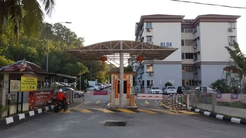 Exterior type: low-rise apartment block; features: main entrance gate with canopy, guard house at entrance, visible parking area, surrounded by greenery, multiple storeys, residential units visible, paved driveway, traffic calming measures with speed bumps; style: modern.