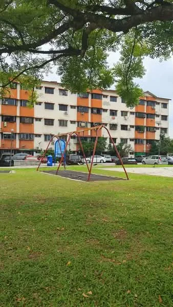 Exterior type: low-rise apartment block; features: orange and white painted facade, 3-storey building, windows with grills, surrounded by grass, playground with swings, parking spaces visible, mature trees providing shade; style: modern.