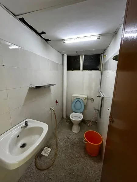 Bathroom features: white tiled walls, grey flooring, single-bowl ceramic sink, chrome faucet, toilet with blue seat, wall-mounted mirror, shower head, window for natural light, orange bucket.