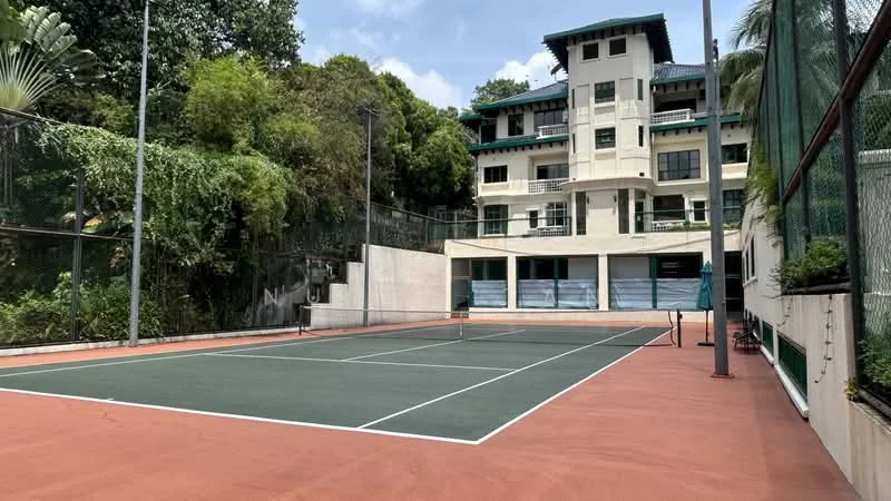 Exterior type: low-rise apartment block; features: tennis court with green surface, surrounded by fencing, landscaped greenery in background, multi-storey building in background, balconies visible on building, paved walkway leading to court; style: modern.