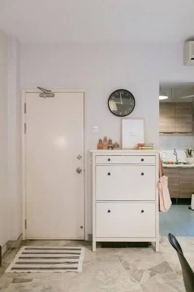 Room features: white painted walls, marble tile flooring, white cabinet with three drawers, wall clock, door with silver handle, rug at entrance, kitchen area visible in background; style: modern.