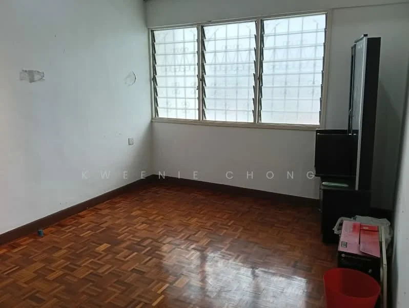 Room features: wooden flooring, white painted walls, large windows with grid pattern, corner TV cabinet, red plastic bin, minimal furniture, natural light from windows.