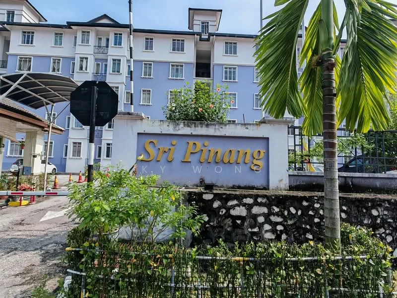 Exterior type: low-rise apartment block; features: blue painted facade, signage with 'Sri Pinang', landscaped garden with plants, stone wall base, paved walkway, parking area, canopy entrance, traffic sign, surrounding greenery; style: modern.