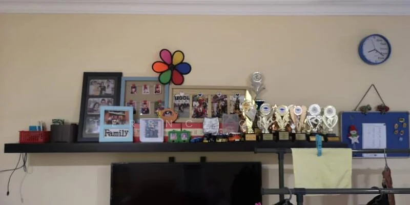 Room features: light yellow painted walls, wooden shelf, family photo frames, colorful flower decoration, trophies on display, blue wall clock, bulletin board with notes, red storage basket, TV console, decorative items; style: modern.
