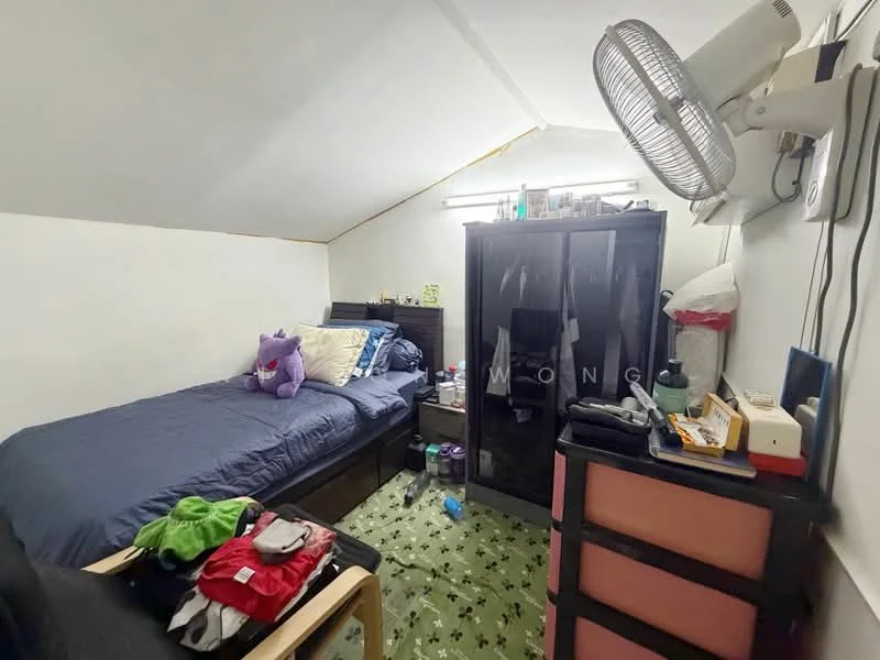 Bedroom features: single bed with blue bedding, upholstered headboard, black wardrobe, wooden bedside table, table lamp, wall-mounted fan, green carpet with pattern, clothes on the floor, decorative plush toy, shelves with items, white walls, sloped ceiling.