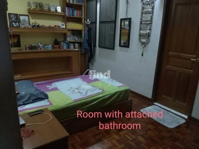 Bedroom features: wooden flooring, yellow painted walls, built-in shelving unit, queen-size bed with green and white bedding, decorative wall hanging, desk with chair, door leading to bathroom, window with curtain, area rug, framed artwork, bookshelves with books.