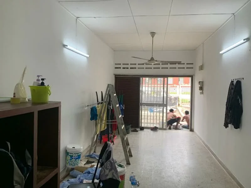 Room features: white painted walls, concrete flooring, ceiling fan, two wall-mounted light fixtures, metal security grille door, wooden door, ladder, cleaning supplies, plastic storage containers, clothes hanging on hooks, water bottle, tools on the floor.
