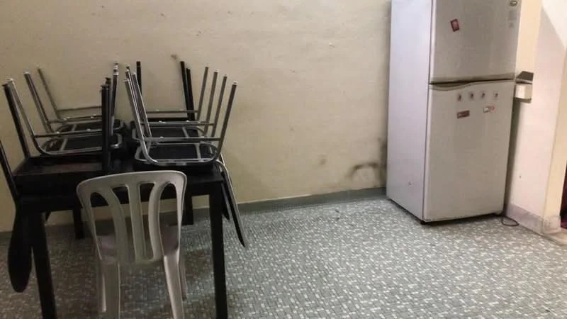 Kitchen features: grey speckled tile flooring, white painted walls, refrigerator, black dining table, stacked chairs, single plastic chair.