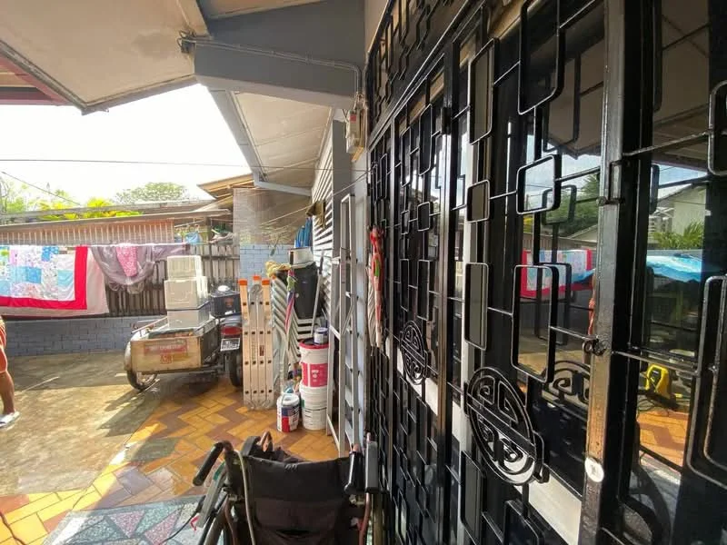 Room features: tiled flooring with geometric pattern, metal security grille door, exposed brick wall, laundry area with hanging clothes, outdoor seating area, storage shelves, bicycle visible, various household items, natural light from outdoor, partially shaded area.